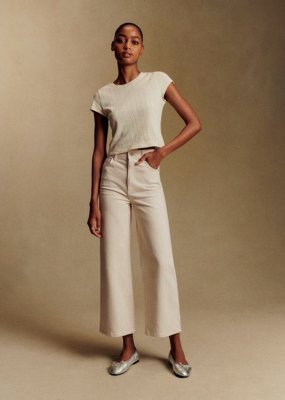 A woman stands against a beige background, wearing a short-sleeved cream top, high-waisted wide-leg cream pants, and silver ballet flats. One hand rests in her pocket.