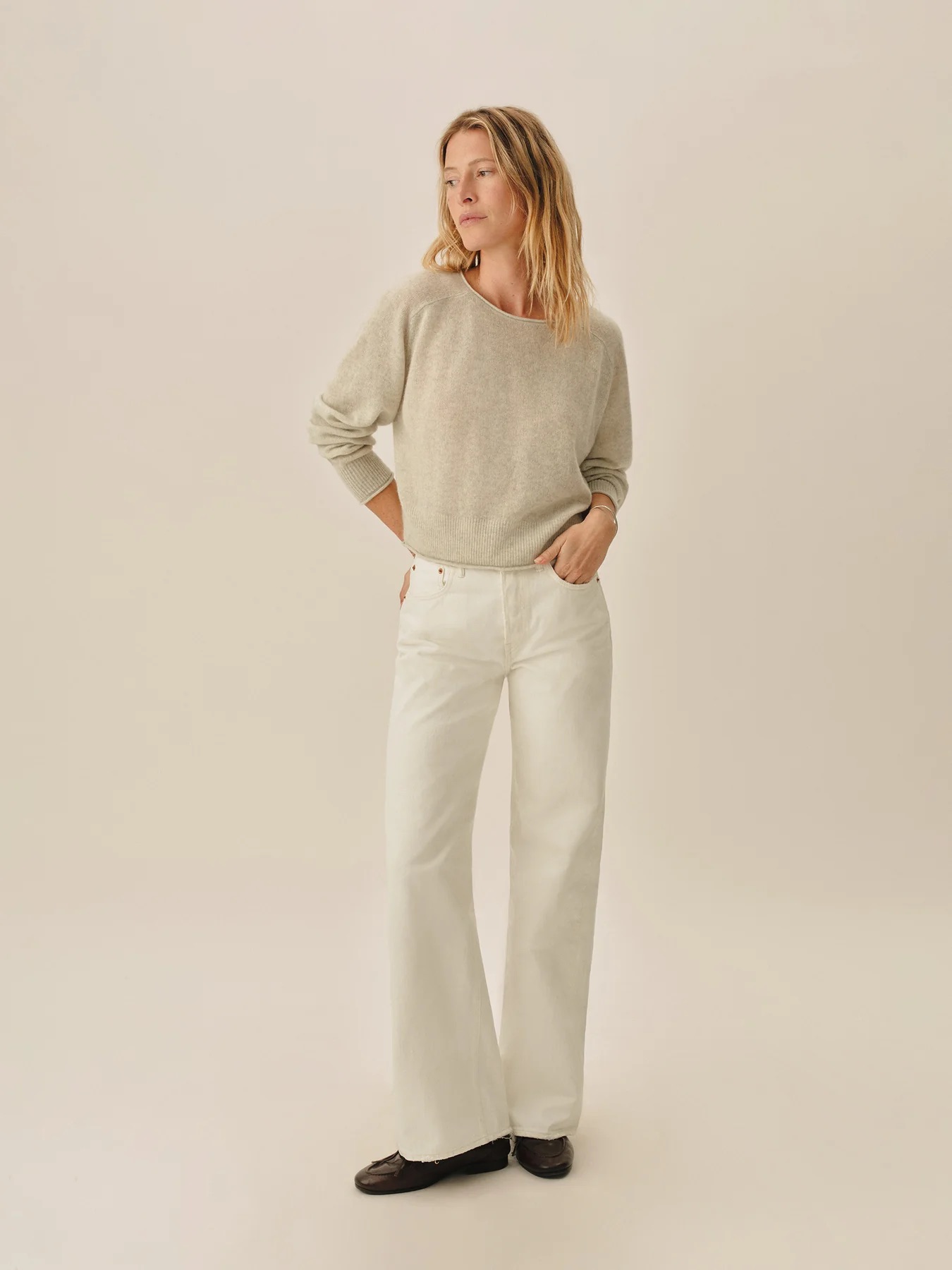A person stands against a plain background wearing a light gray sweater, white wide-leg pants, and dark shoes, with hands in pockets and looking to the side.
