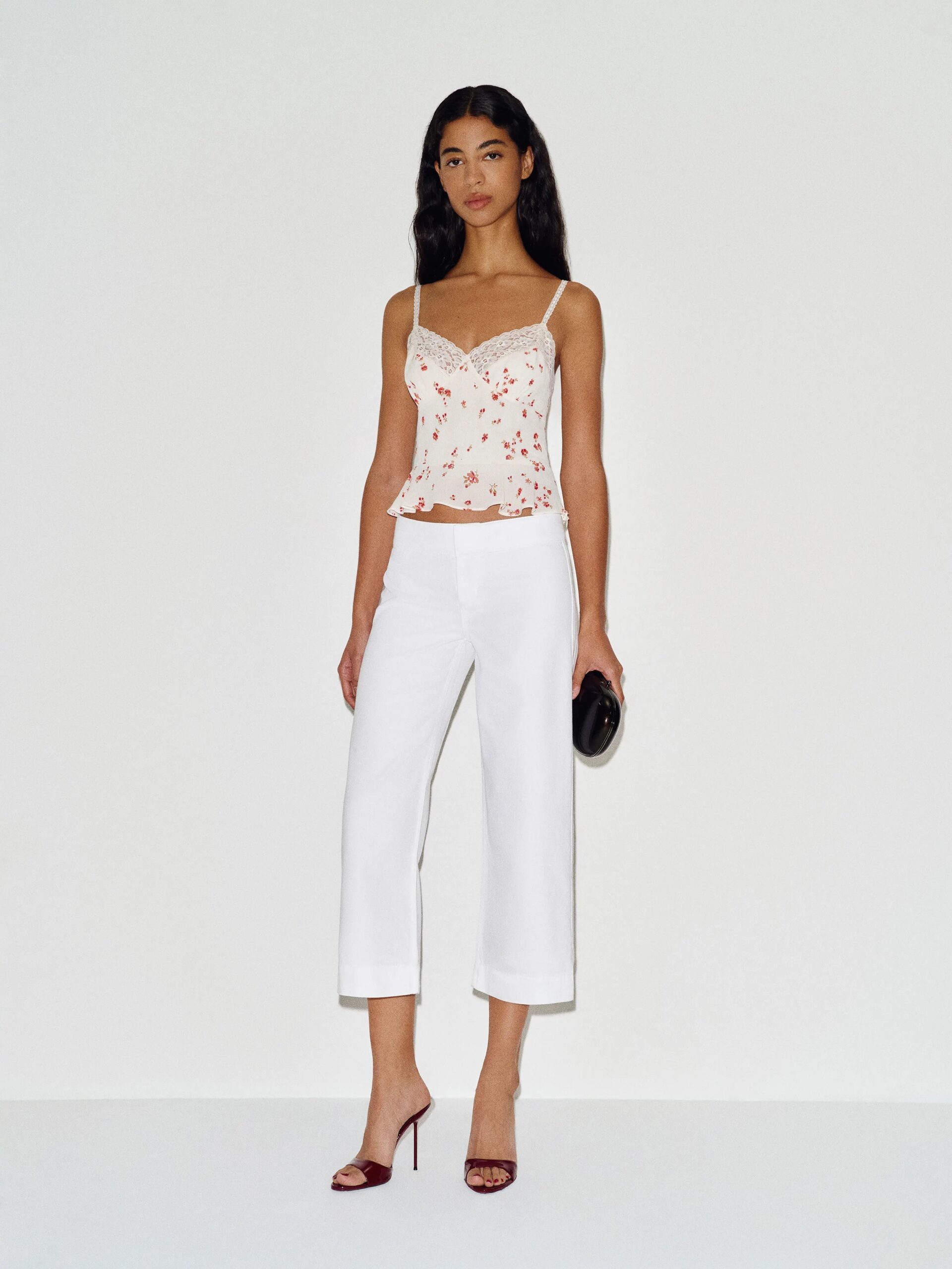 A woman stands against a plain white background wearing a floral camisole top, white cropped pants, maroon heels, and holding a black clutch.