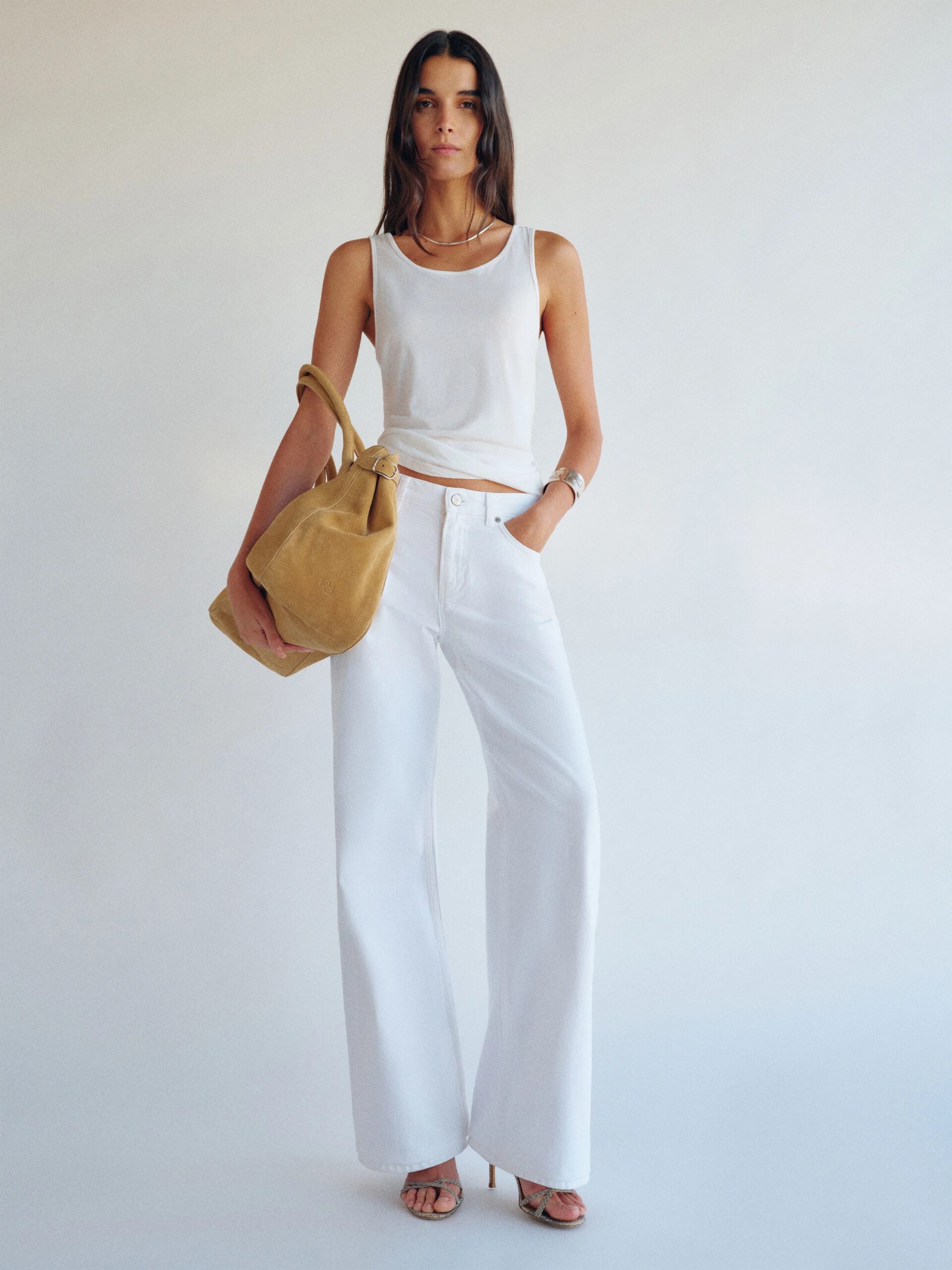 Woman stands against a plain background wearing a white sleeveless top, white wide-leg pants, heels, and carrying a large tan bag on her shoulder.