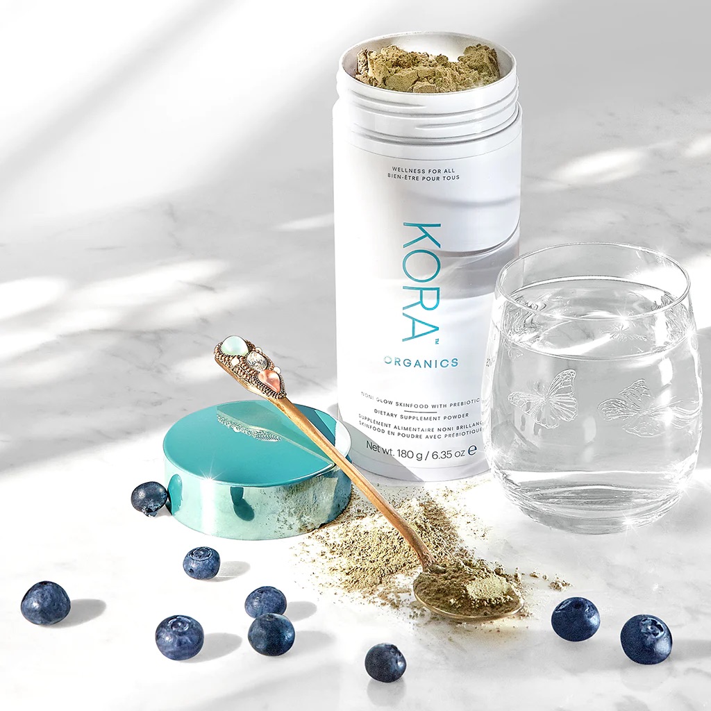 An open container of Kora Organics Glow Skinfood next to a spoon filled with the powder, a glass of water, and scattered blueberries.