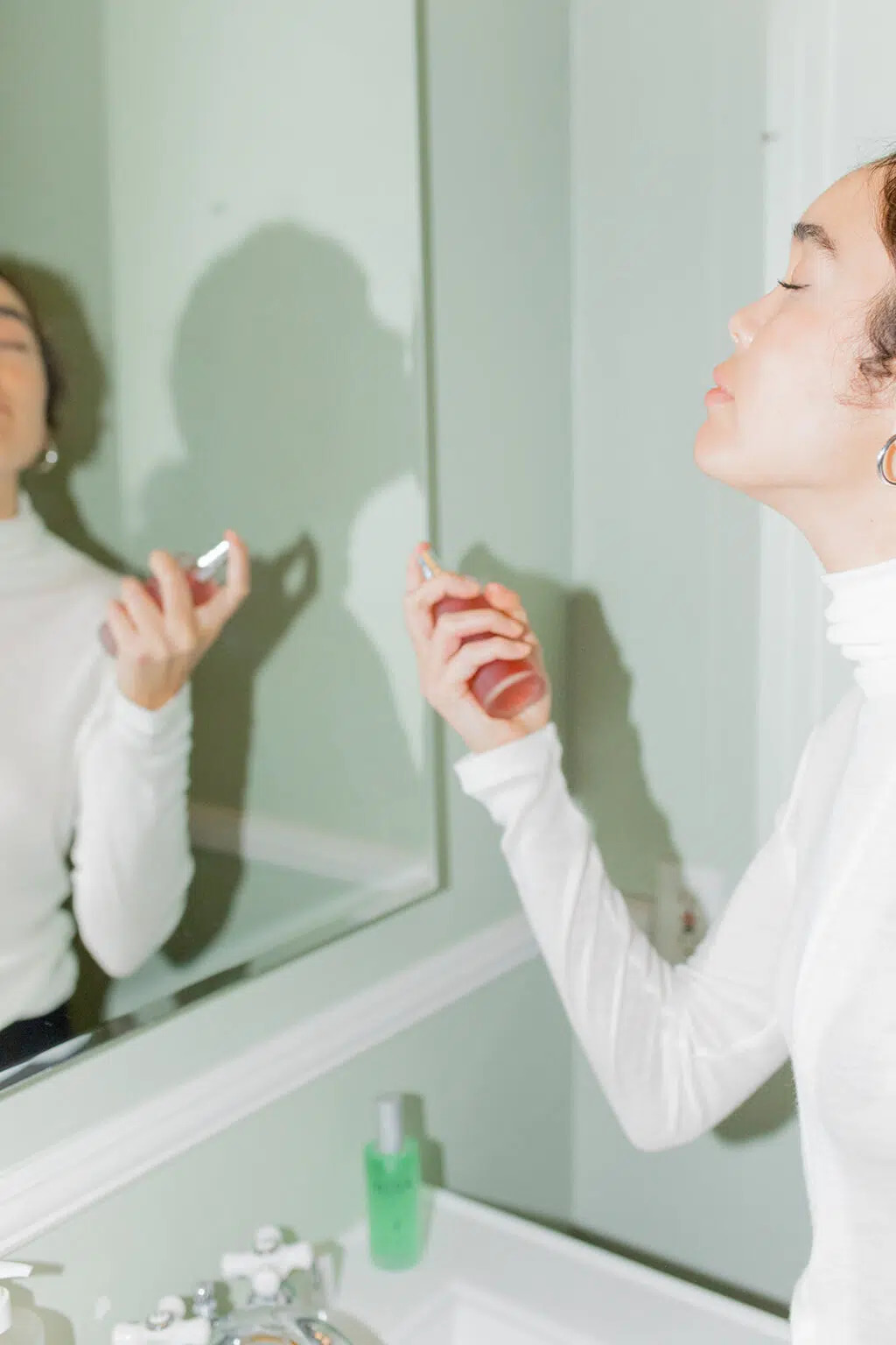 A woman facing a mirror in a restroom with green walls with her eyes closed as she sprays witch hazel toner on her face.