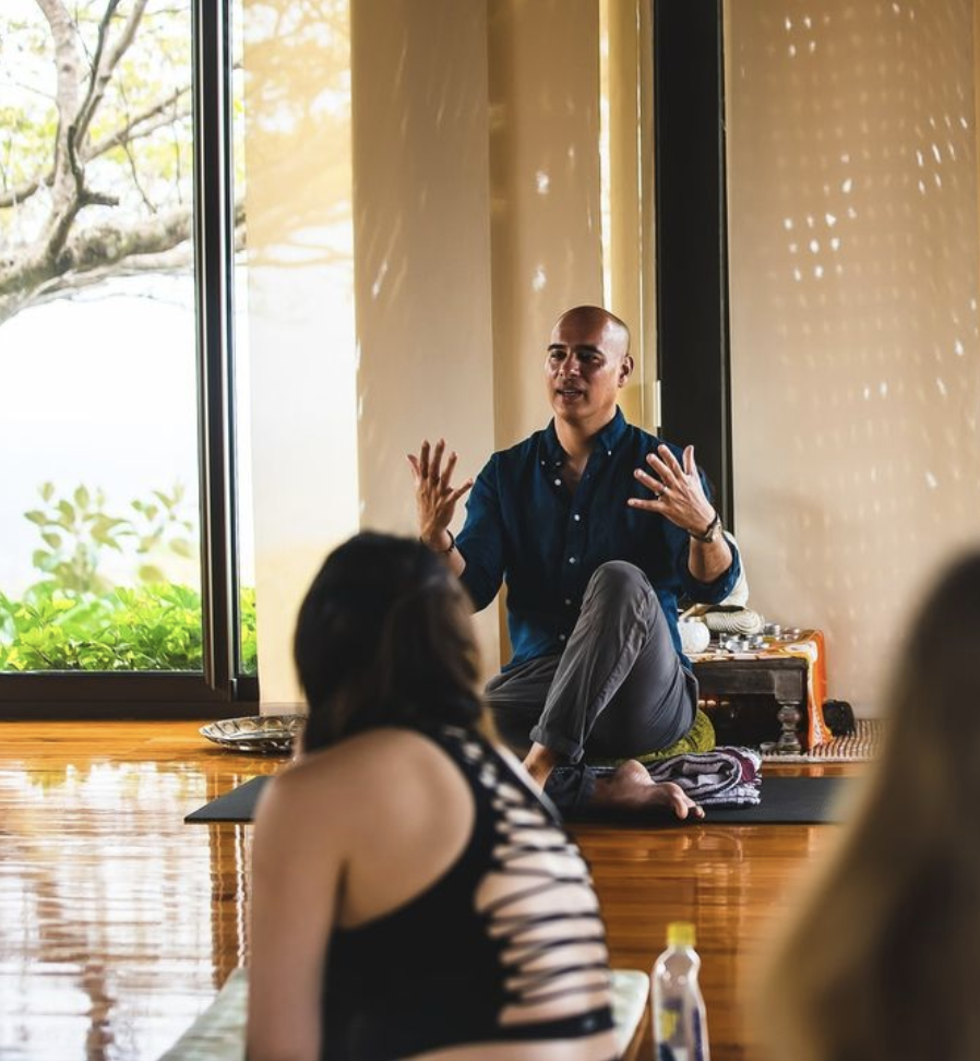 A adult man sits on a yoga mat and gestures to a class. 