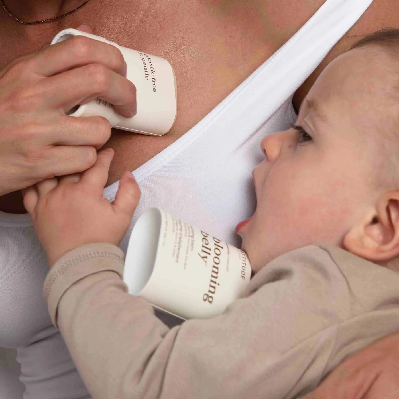 An adult holds a small container near their chest while a baby, lying close, reaches for another similar container, both featuring printed text.