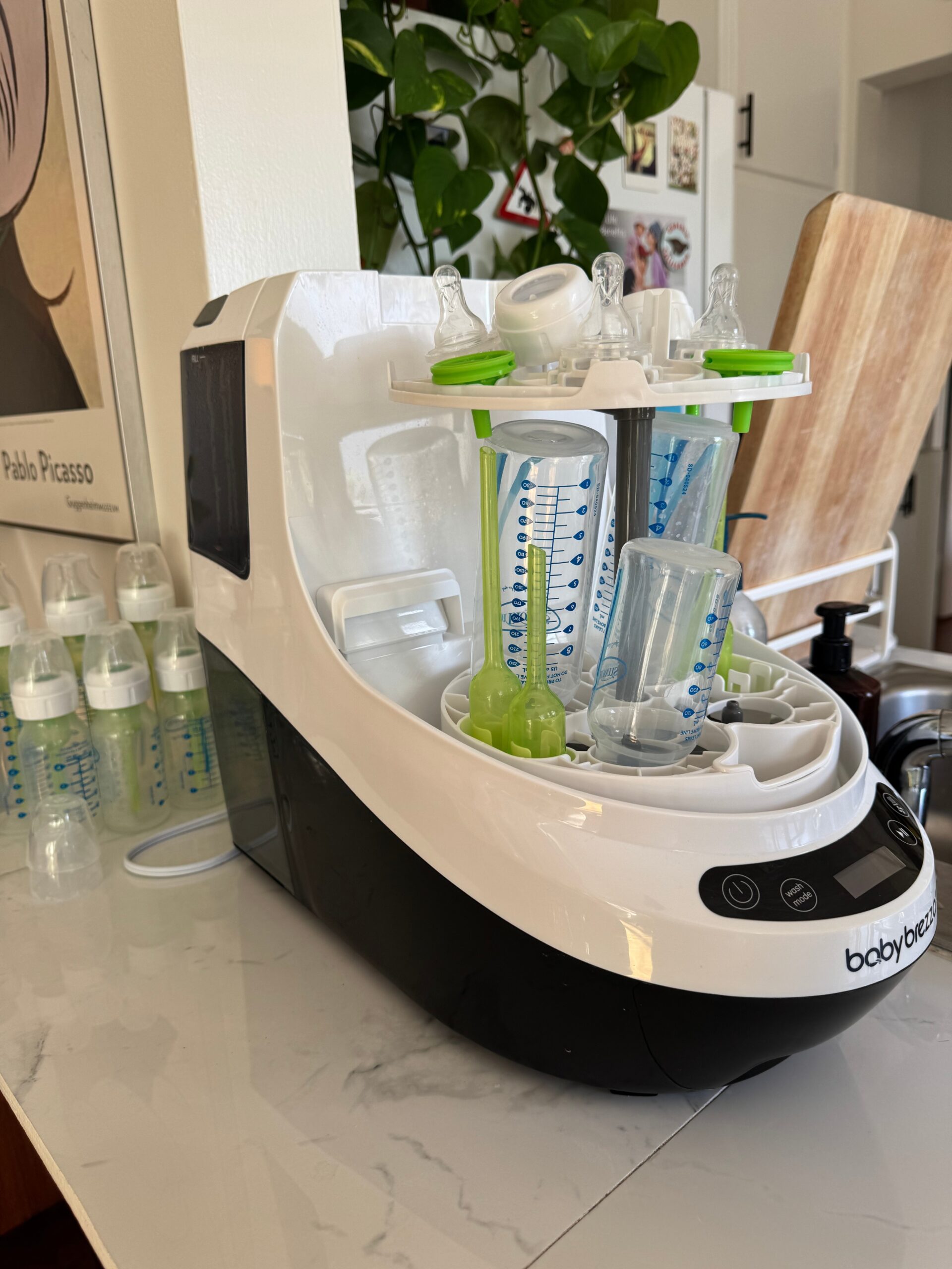 A baby bottle sterilizer sits on a kitchen counter with several clean bottles and accessories inside and more bottles drying in the background.