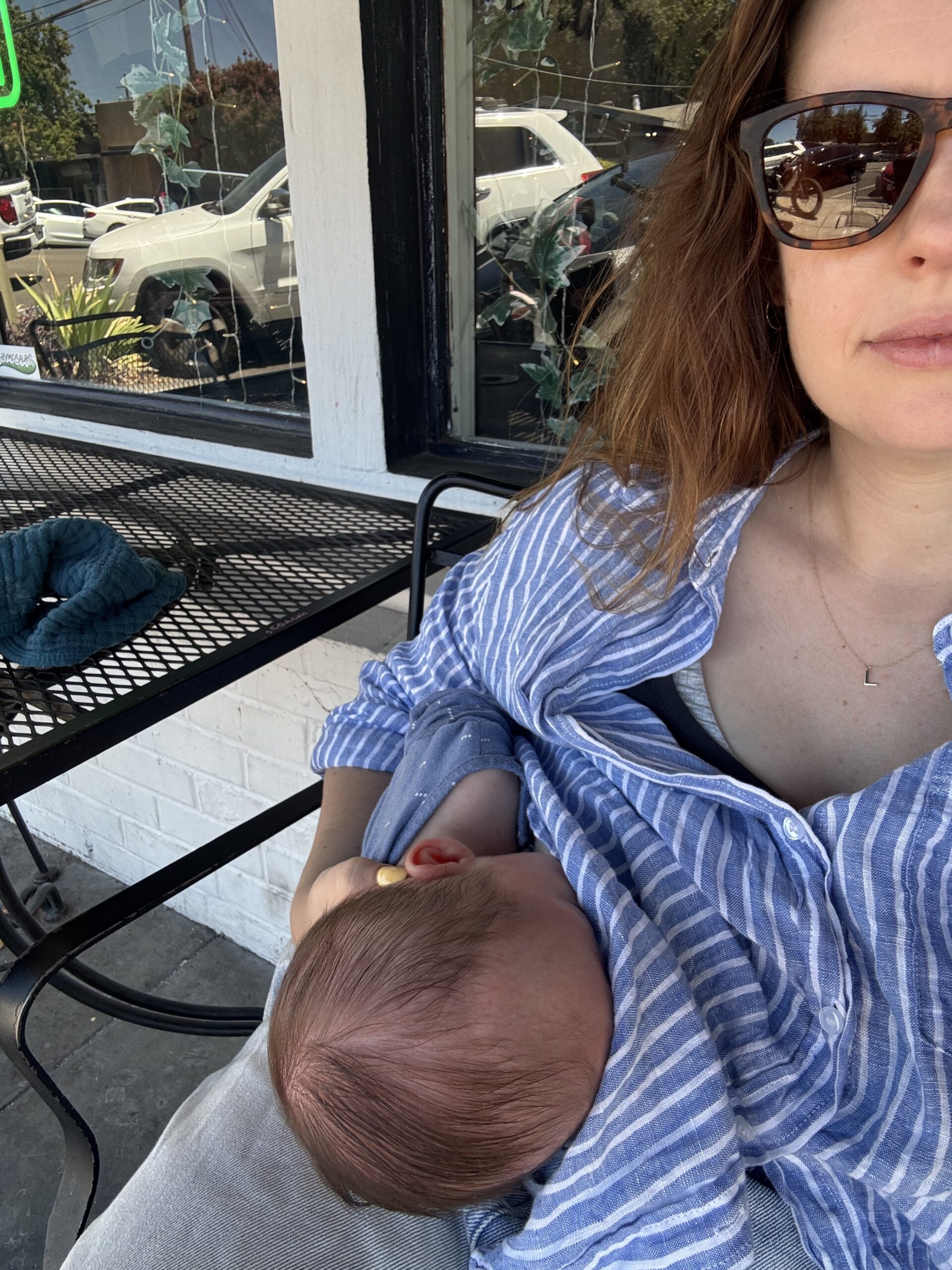 Woman wearing sunglasses and a striped shirt is breastfeeding a baby while sitting outside at a cafe with a metal table and plants visible in the background.
