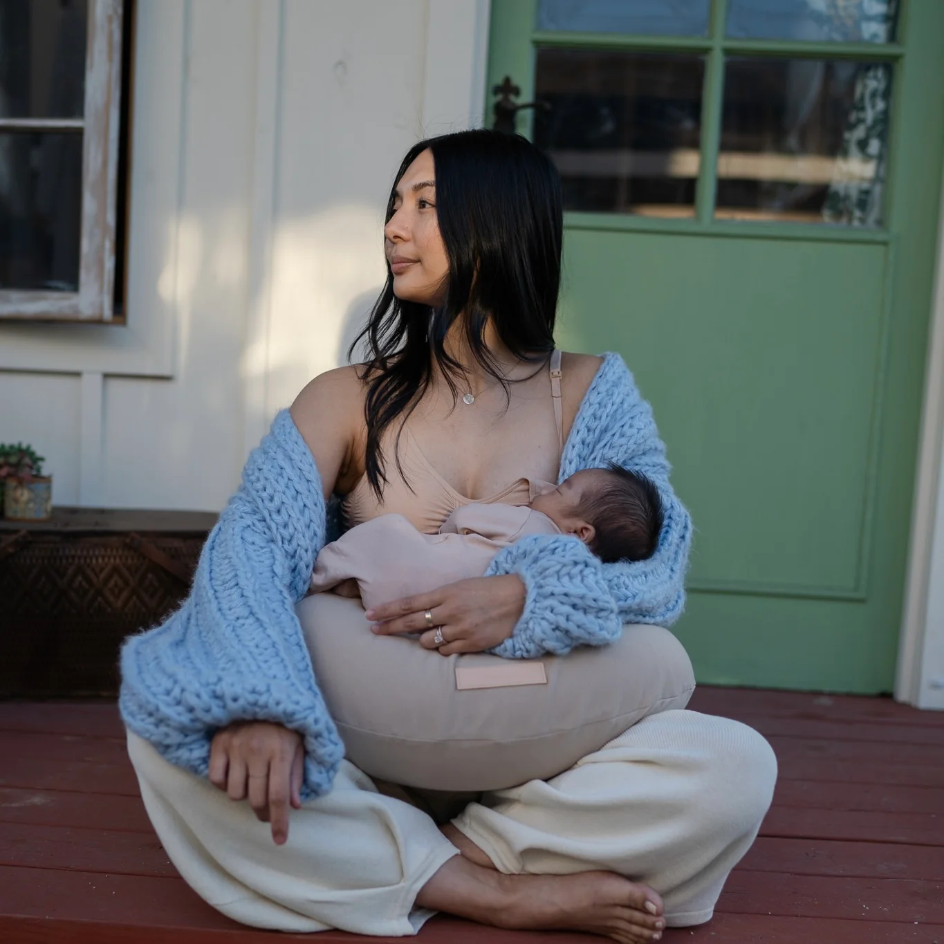 A woman sits cross-legged on a porch, wearing a blue sweater and light pants, breastfeeding a baby with the aid of a support pillow.