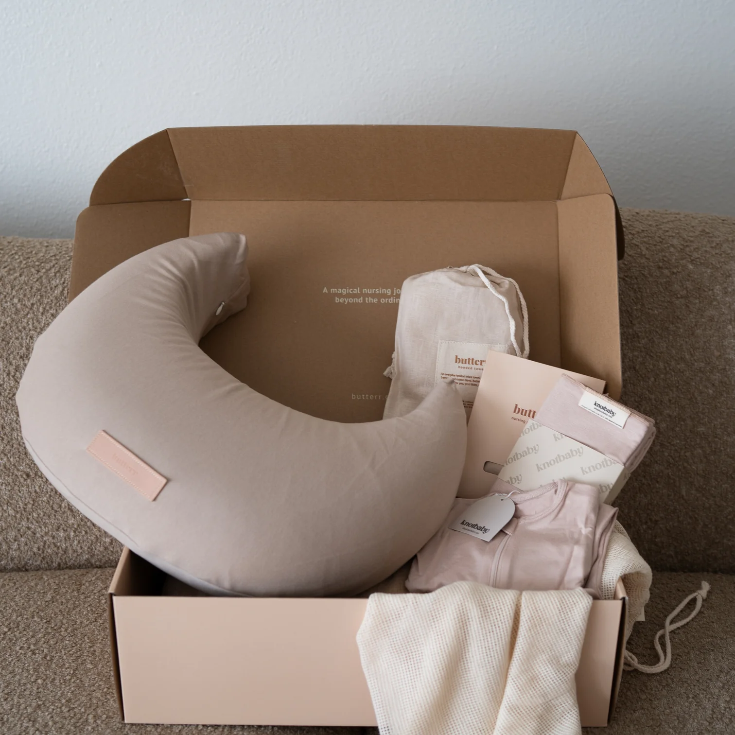 A gift box contains a beige nursing pillow, a pouch, folded fabric items, and informational cards, all arranged neatly on a beige sofa.