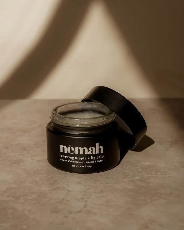 An open jar of Nomah renewing nipple and lip balm sits on a beige surface in natural light, with its lid resting beside it.