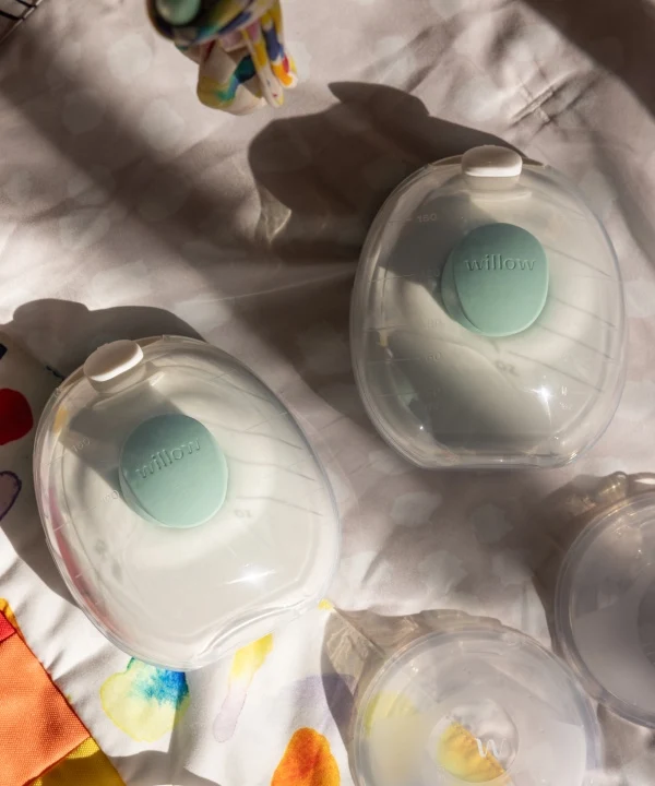 Two wireless breast pumps labeled “Willow” rest on a white patterned surface with colorful fabric and lids nearby.