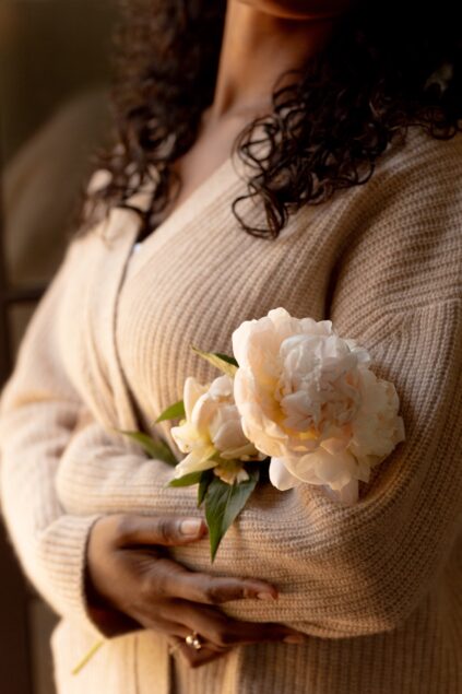 A person in a beige knit sweater holds a stem with two white peony flowers, with their arms crossed over their chest.