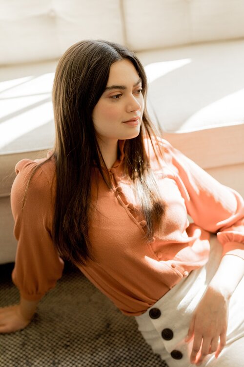 A model in a coral long sleeve blouse and high waisted white pants seated against a ledge.