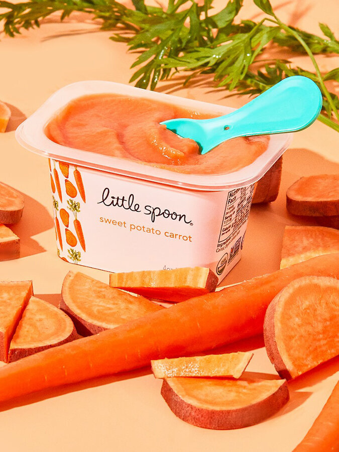 A container of sweet potato carrot puree with a spoon sitting in it, surrounded by slices of sweet potatoes and whole carrots.