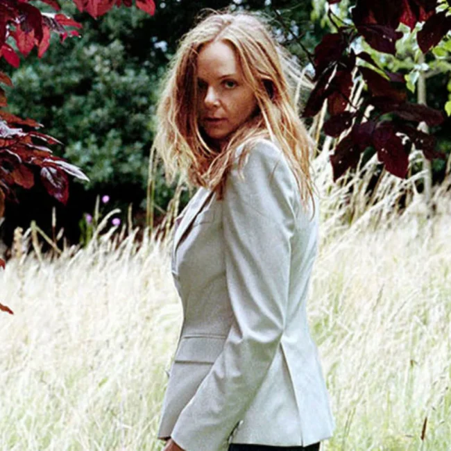 A person with long hair wearing a light-colored blazer stands outdoors in a grassy field, framed by red and green foliage.