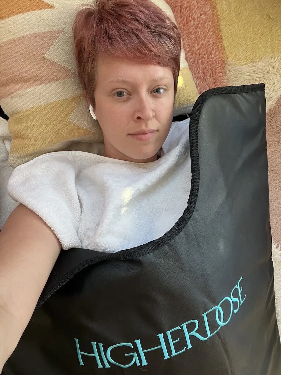 Person with short pink hair lies on a pillow, wearing a white top and AirPods, partially covered by a black mat labeled "HIGHERDOSE.