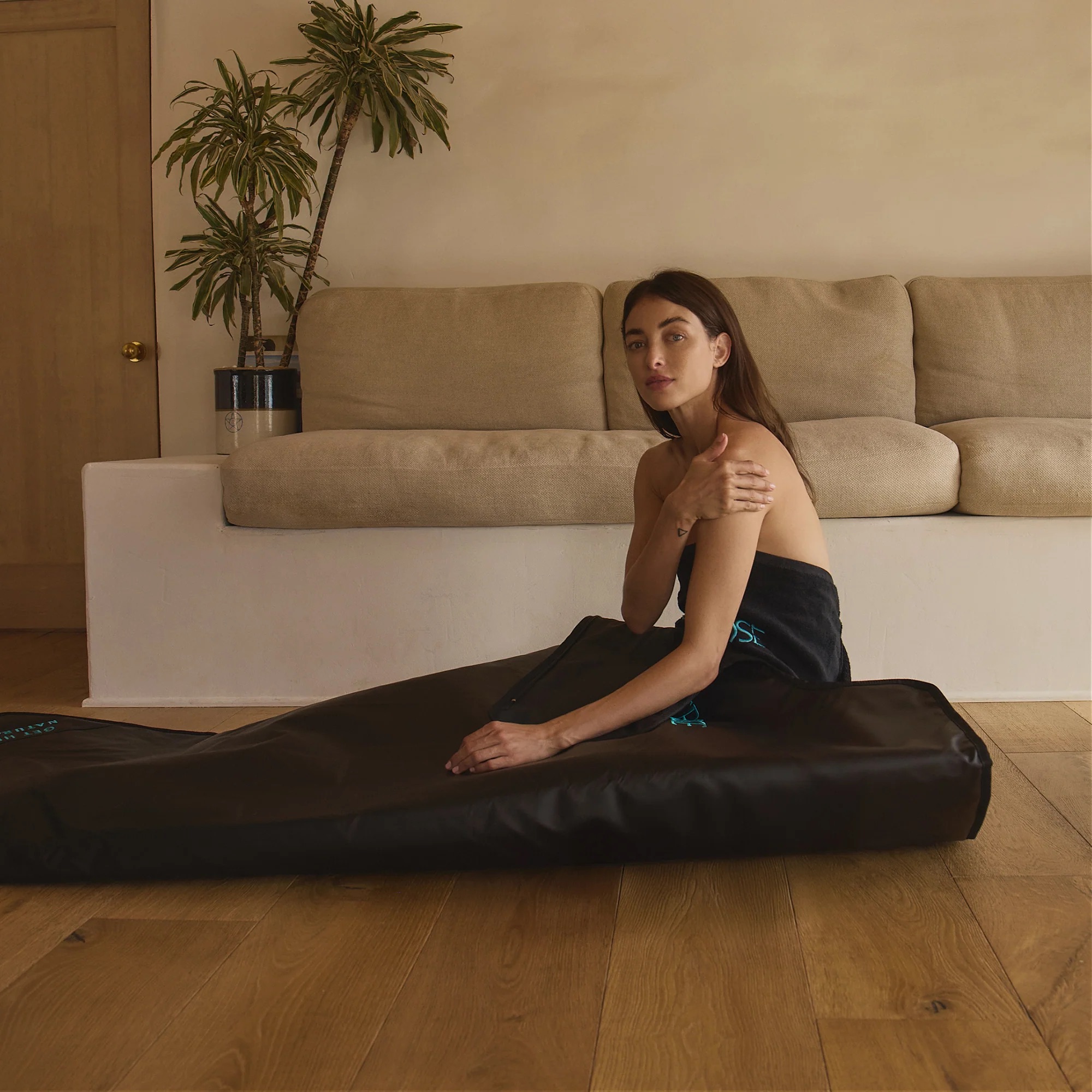 A woman sits wrapped in a black towel next to a surfboard bag in a room with a couch and plants.