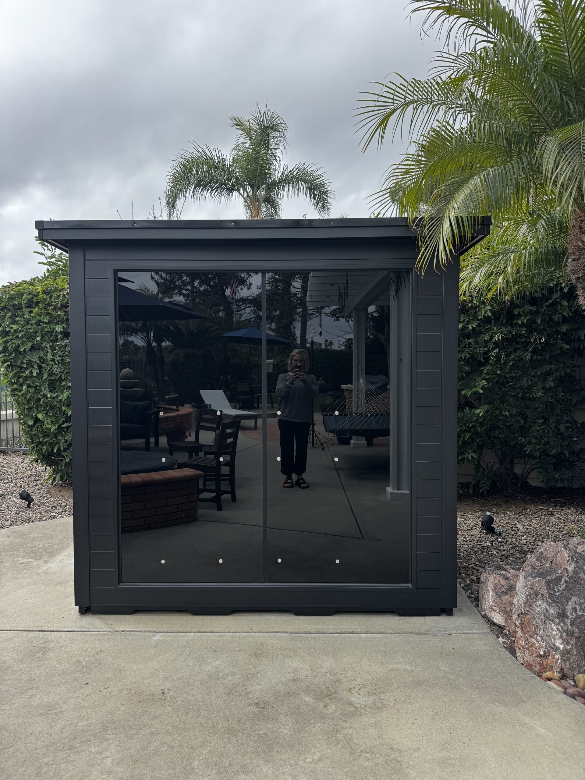 A person is reflected in the glass doors of a small, modern garden shed set on a concrete patio, surrounded by palm trees and outdoor furniture.