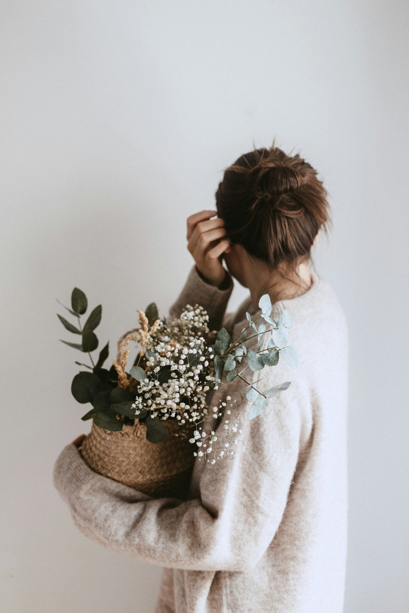 A person in a beige sweater holds a woven basket filled with eucalyptus and baby’s breath.