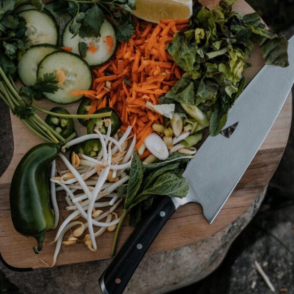 A chopping board with fresh vegetables including cucumbers, carrots, lettuce, and a jalapeño beside a chef's knife.