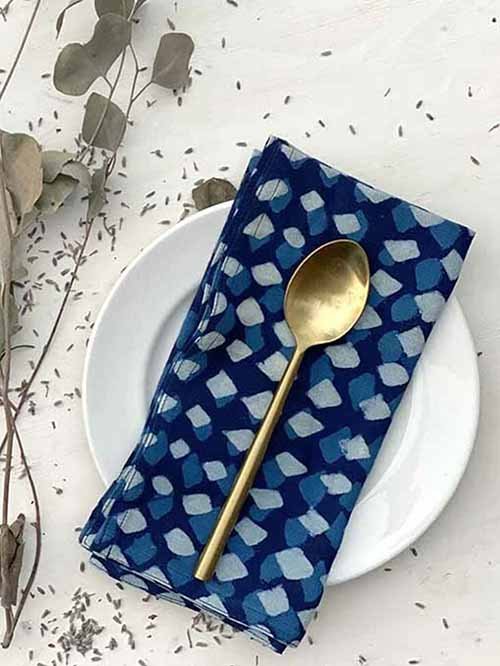 Cloth Napkins: Ichcha's blue block-printed organic cotton napkin lays on a white salad plate on a white tablecloth surrounded by greens, and the napkin is topped with a gold spoon.