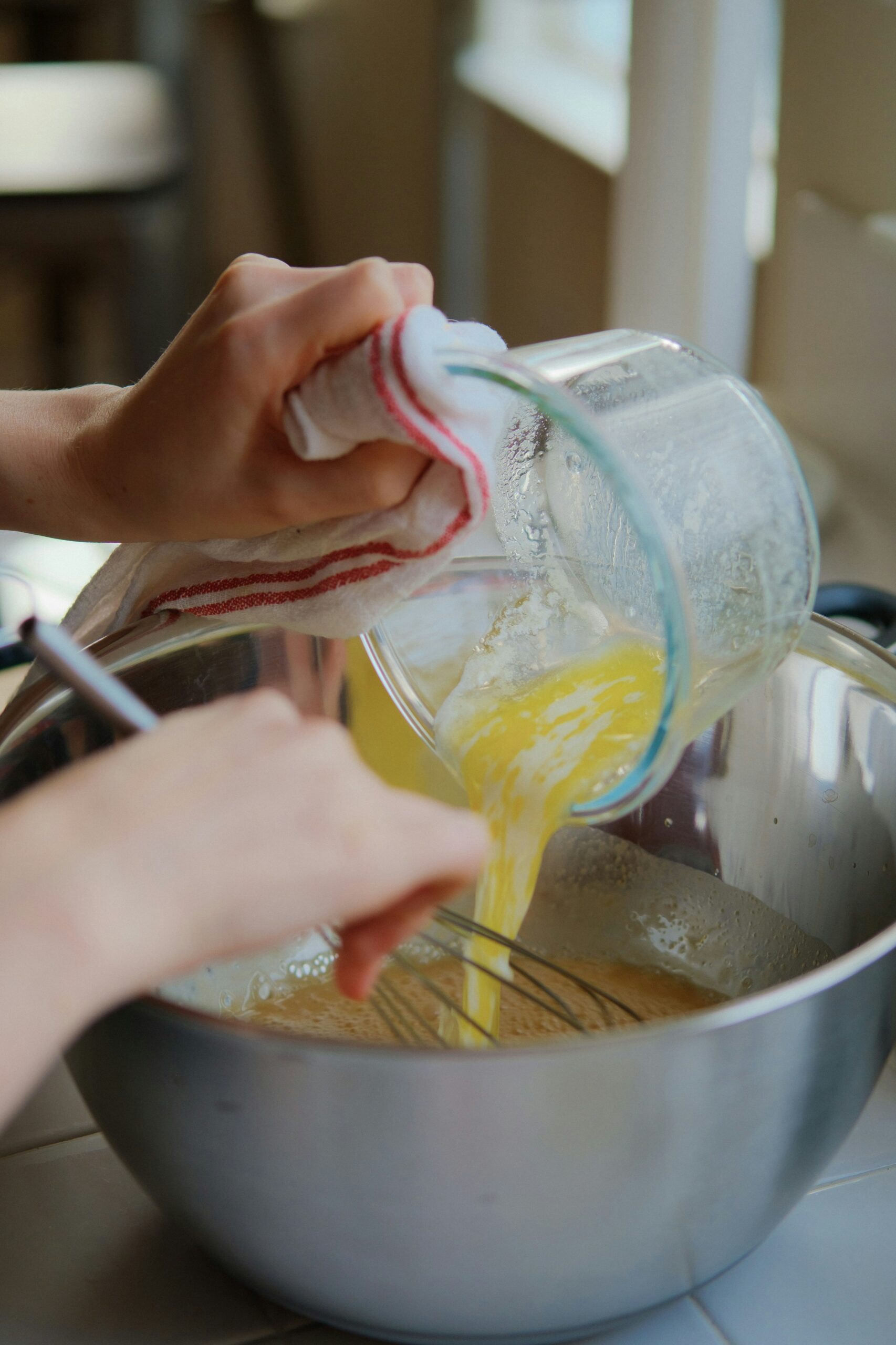 Hands pouring melted butter from a glass measuring cup into a metal mixing bowl while whisking.