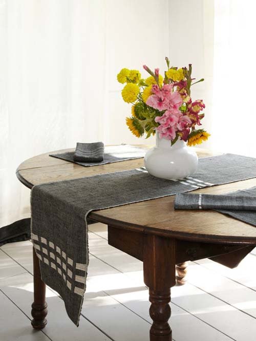 Tablecloths & Runners: Bole Road Textile's Allen Table Runner in the onyx pattern. The runner, matching placemats, and napkins are all on a brown wooden table. A rounded vase with flowers sits atop the runner.