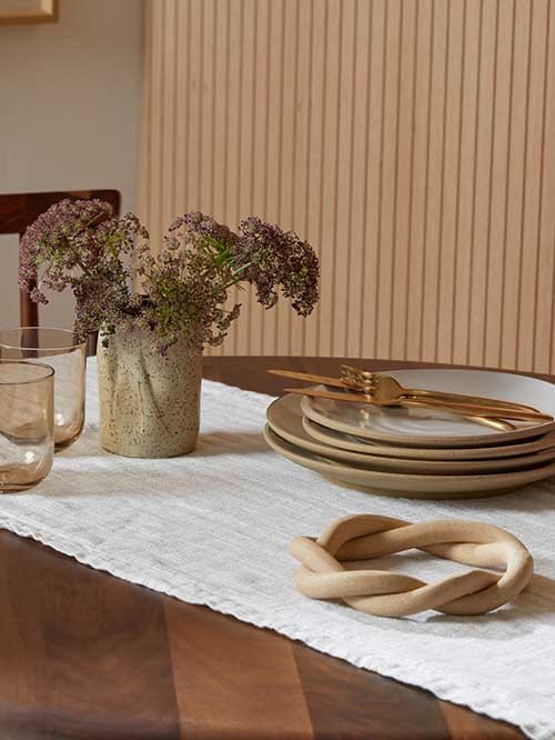 Tablecloths & Runners: Parachute's Linen Waffle Tabletop Runner in cream, laid across a wood table. On top of the runner is two glasses, a vase with flowers, a set of dinner plates, and a knotted trivet.