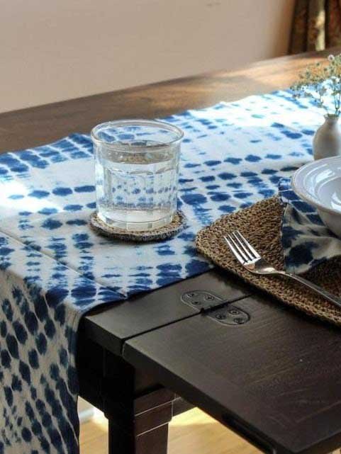 Tablecloths & Runners: Terraklay's Geetha Shibori Tie-Dye Runner lays on a dark brown table, with a glass and coaster on top of the runner. The runner is blue and white tie-dye in shibori patterns.