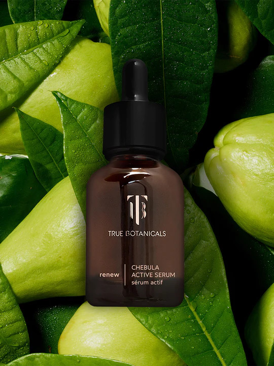 A dropper bottle of True Botanicals Chebula Active Serum with green leaves and green fruits displayed behind it.