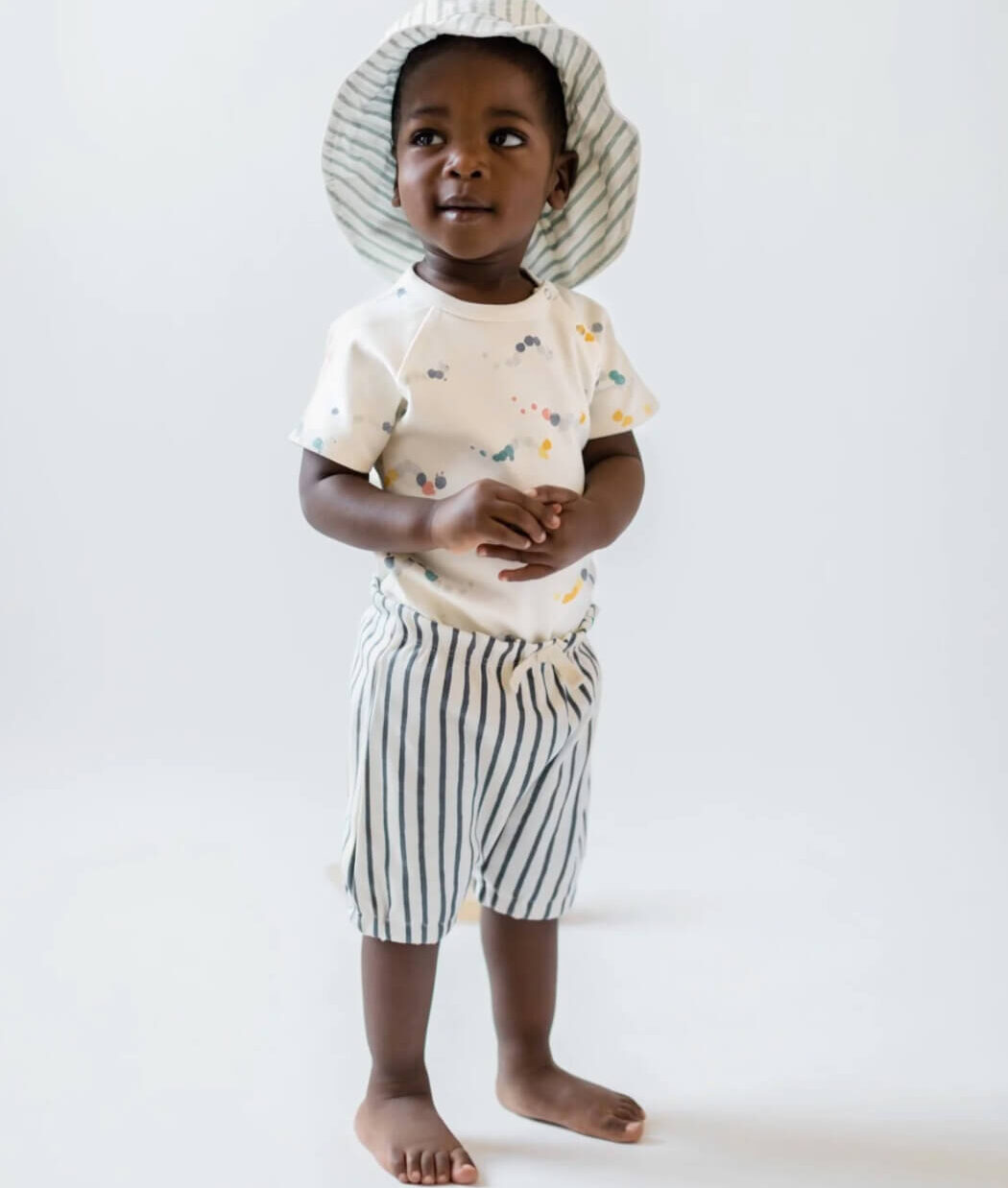 A baby wearing a striped bucket hat, an abstract print shirt and striped shorts from Pehr.