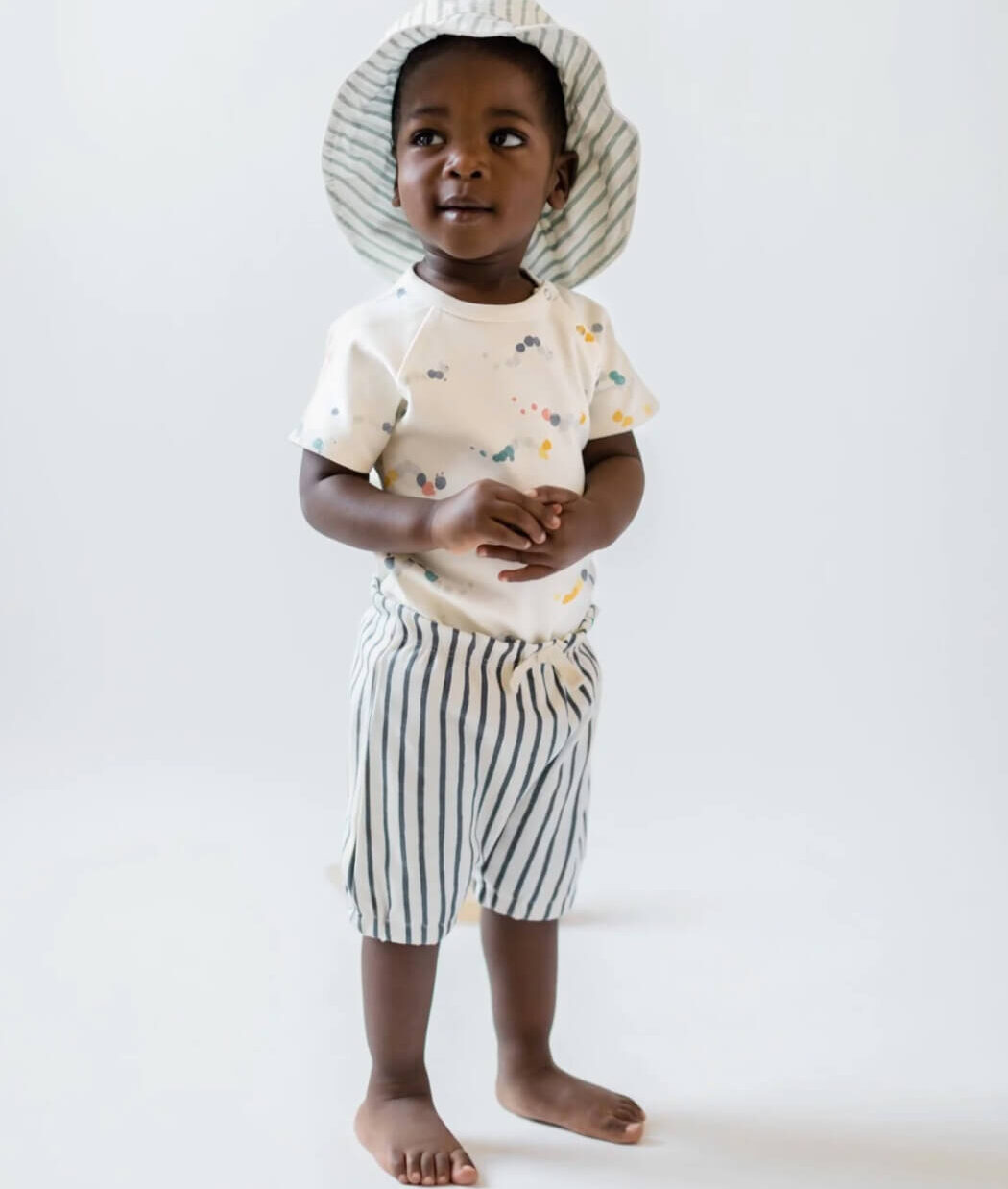 A baby wearing a striped bucket hat, an abstract print shirt and striped shorts from Pehr. 