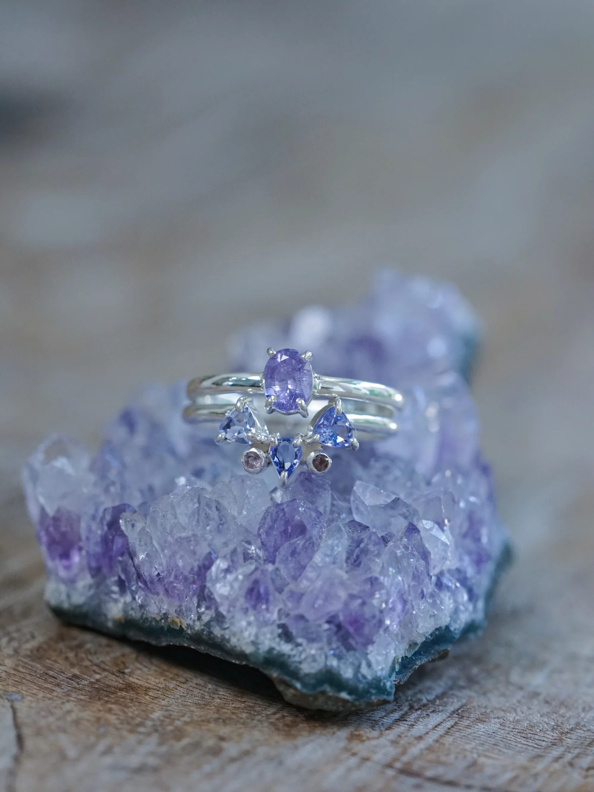 Silver ring with a central purple gemstone flanked by clear stones, displayed on an amethyst crystal cluster.