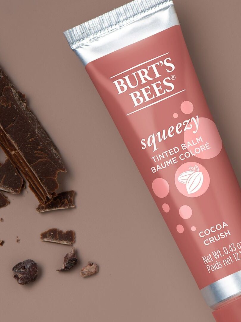 A closeup of Burt's Bees Squeezy Tinted Balm with broken chocolate to the left of the product.