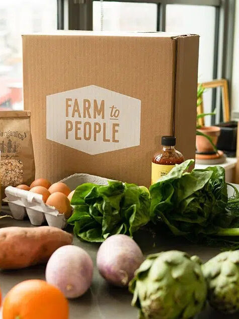 A cardboard box that says Farm to People with vegetables and eggs on a table.