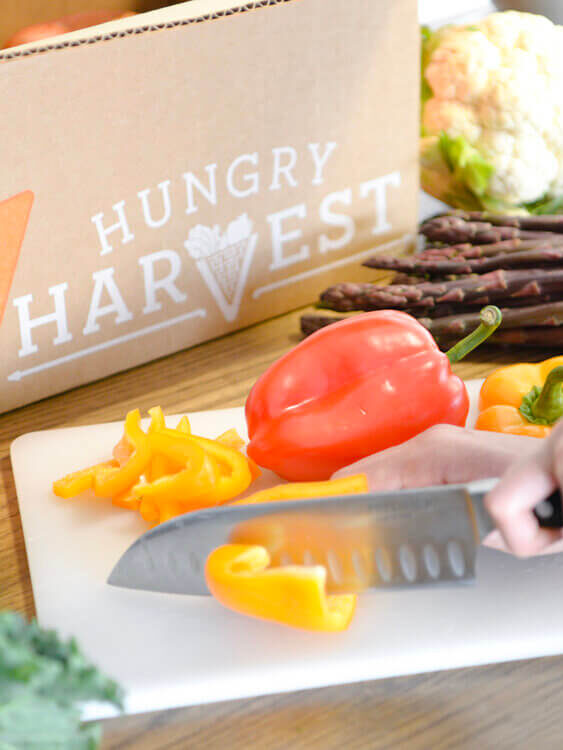 A box of Hungry Harvest sits next to a cutting board where someone cuts a yellow pepper.