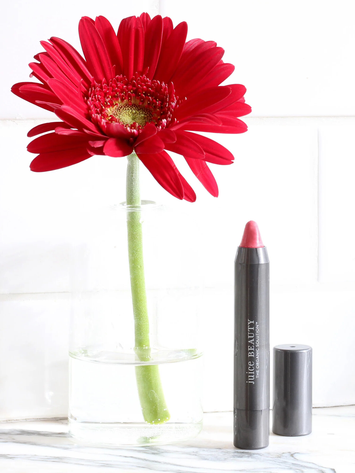 A Juice Beauty jumbo Lip Pencil with a red flower in a clear vase to the left of the pencil.