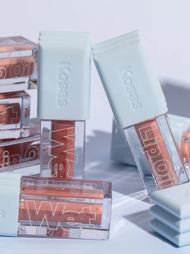 Kosas Lip Oil's in piles and leaning next to each other in front of a baby blue background.