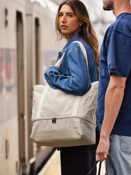 A model carrying a carry-on tote waits for a train. 