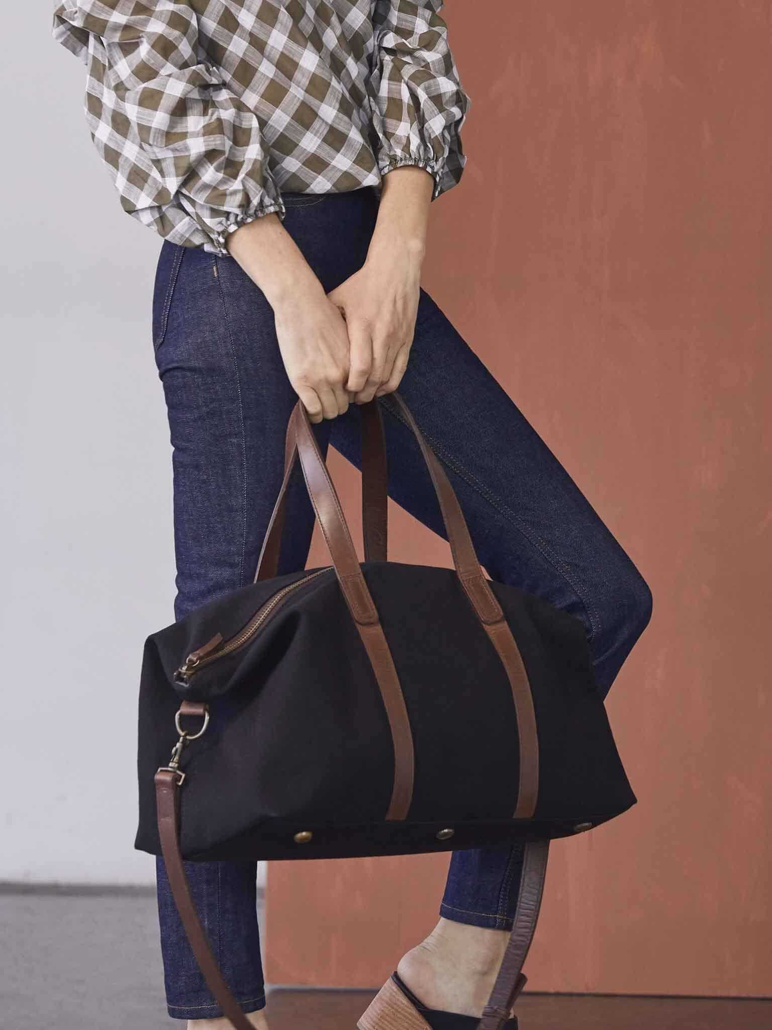 A woman holds a black and brown canvas weekender bag.