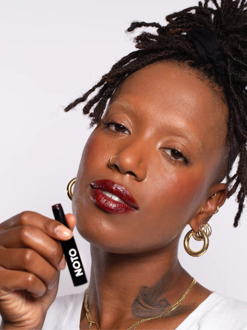 A woman with long locks in an updo and multiple chunky gold hoop earrings wearing Noto's multi-use balm on her lips and holding up the product in her hand.