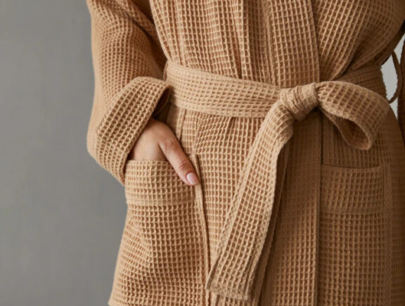 A closeup of the waist area of a person wearing a Coyuchi beige waffle knit robe, with their hand in the pocket.