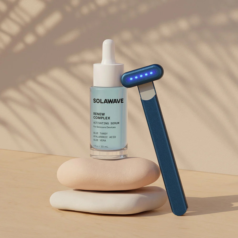 A blue Solawave wand is propped against a serum bottle sitting on a pair of smooth rocks.