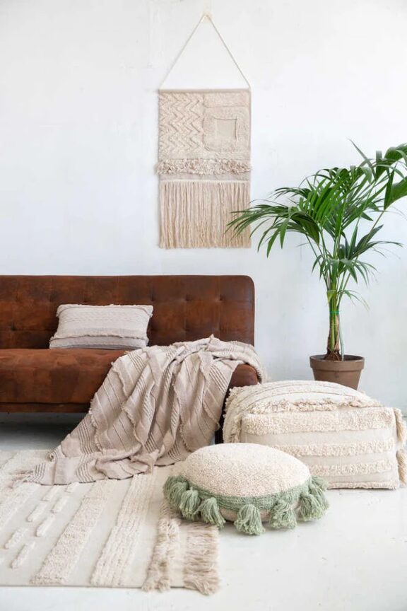 A cozy living room with a leather sofa, decorative macrame wall hanging, floor cushions, a textured rug, and a potted plant.