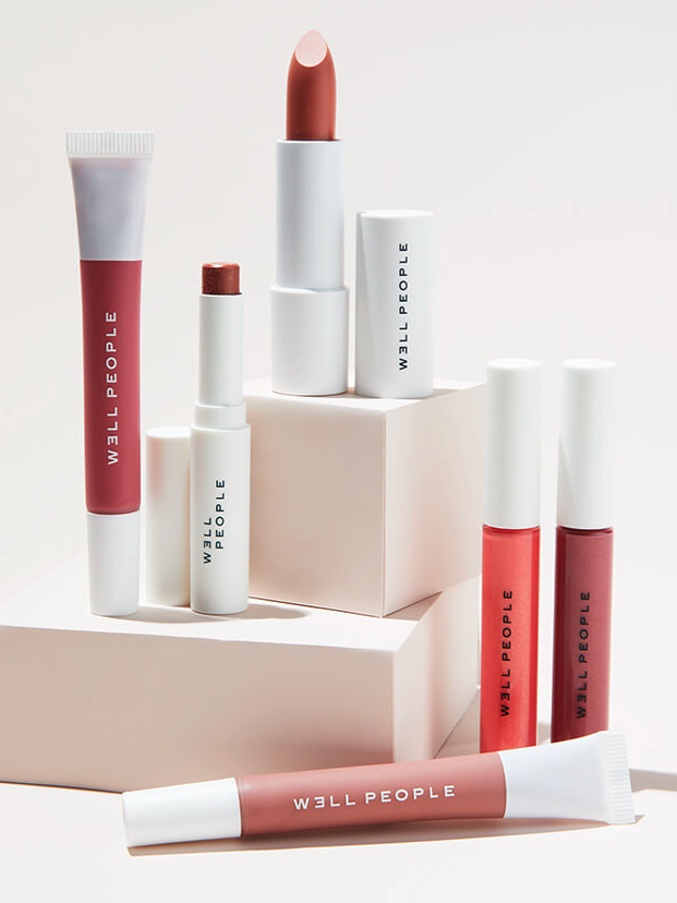 Well People's lip products in white containers displayed in an array, on or in front of white blocks.