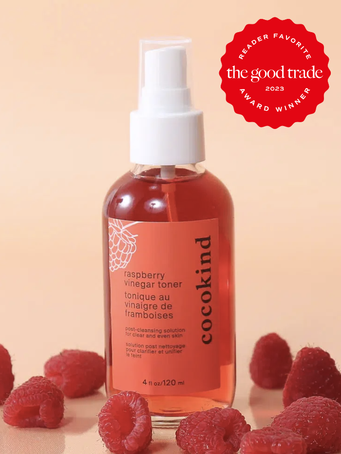 A spray bottle of cocokind's raspberry vinegar toner displayed in front of a pale pink beige background with raspberries on the bottom. The Good Trade's Reader Favorite award sticker in red is on the top right.