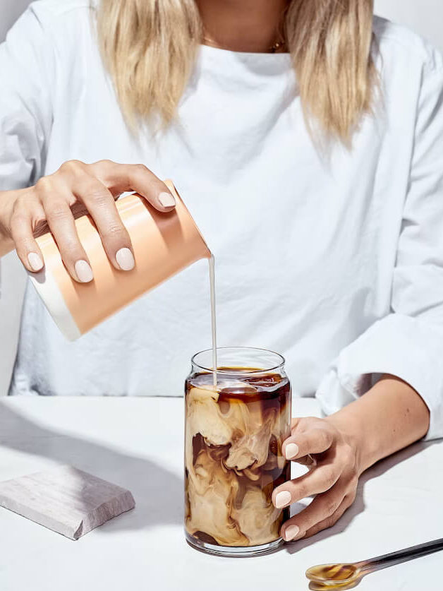 A woman pours creamer from a beaker into a tall glass of Four Sigmatic coffee alternative.