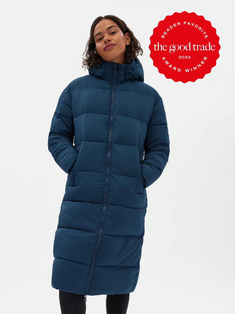 A model wearing a long blue recycled puffer coat by Girlfriend Collective. A red Reader Favorite Award 2023 sticker by The Good Trade is on the top right hand corner.