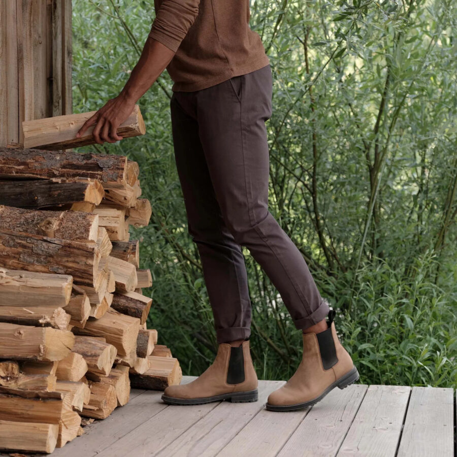 A model waist-down. wearing a brown long sleeve sweater, dark brown pants and light brown Nisolo boots while picking up a log from a pile.