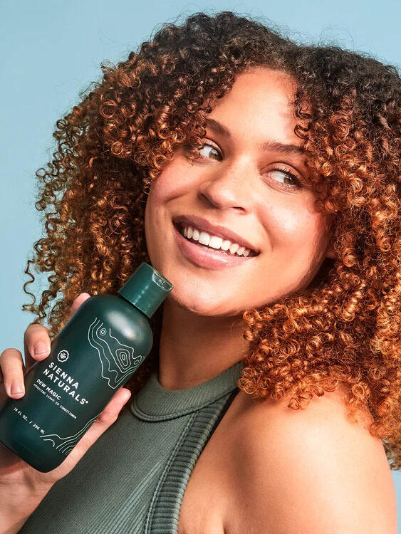 A model with curly hair holds a Sienna Naturals product hear her face.