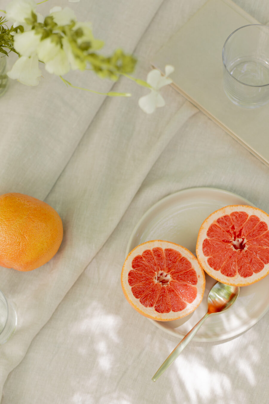 A halved grapefruit on a plate with a spoon, a whole grapefruit nearby, and clear glasses on a light fabric tablecloth, with flowers and a book in the background.